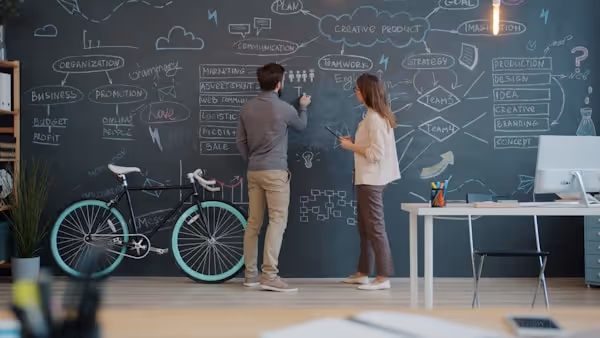 Coworkers discussing strategy at a chalkboard in an open office